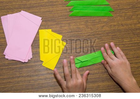 Boy Is Making Origami From Paper Of Green, Pink And Yellow Colors On The Desk Background