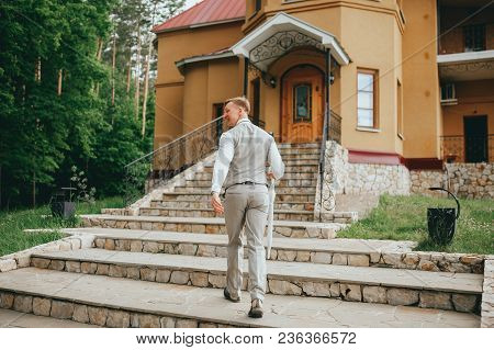 Groom In The Stylish Wedding Suit Going To The Bride With A Bouquet Of Flowers. Back View