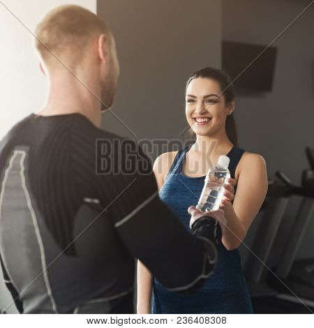 Fitness Couple Having Rest And Talking, Sharing Water At Gym. Smiling Woman Giving Plastic Bottle To