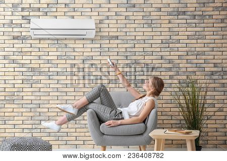 Young woman switching on air conditioner while sitting in armchair at home