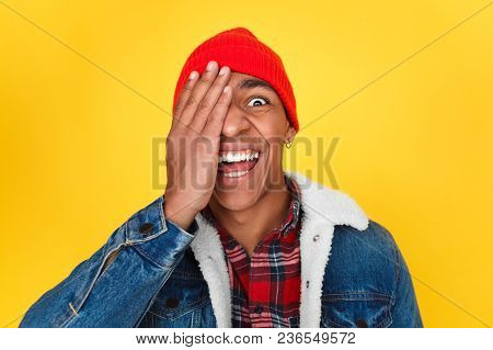 Black Stylish Man Covering One Eye And Looking Crazily And Cheerfully At Camera On Yellow Backdrop.