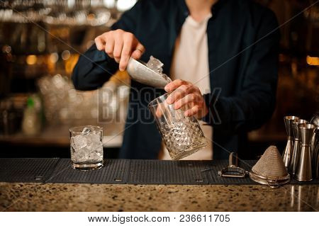 Bartender Putting An Ice Cubes Into An Empty Glass On The Bar Counter On The Blurred Background