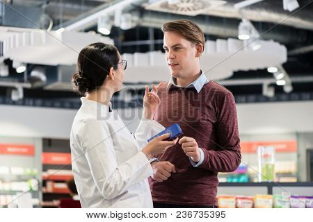 Experienced female pharmacist checking the indications and contraindications of a new medicine next to a young male customer in a modern pharmacy
