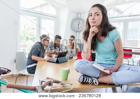 Portrait of a beautiful young woman with cool casual clothes smiling while sitting on a desk during work in a modern co-working space
