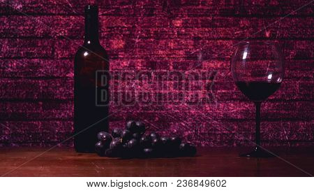On The Table, A Crystal Glass, A Bottle Of Red Wine, Purple Grapes, A Redwood Background.