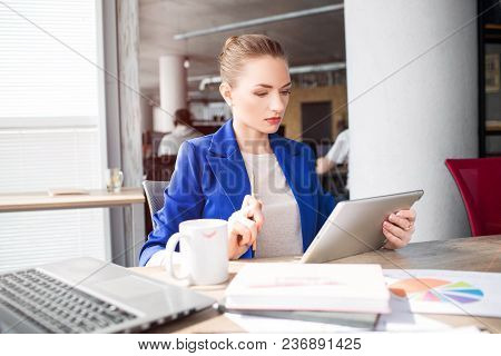 Smart And Good-looking Woman Is Sitting On The Table And Looking To The Tablet. She Is Studying. Gir