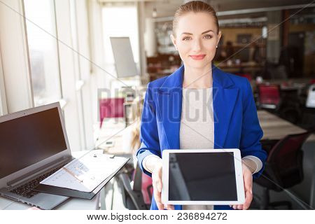 Nice And Cheerful Person Is Standing In An Office Room And Holding A Tablet. She Is Showing A Dark S