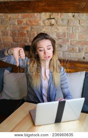 Young Woman Sitting In Cozy Coffee House Near Wooden Table With Glass Cup Of Cappuccino And Using La
