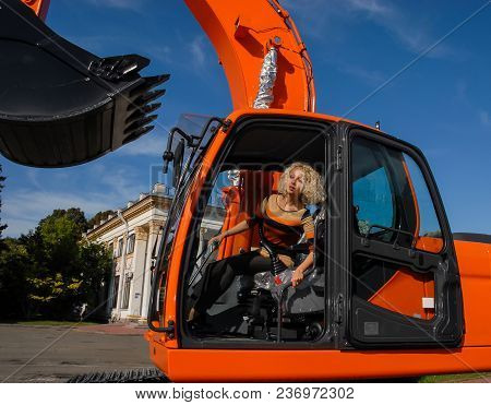 A Young Blond Hair Woman Sits In A Cabin Of A New Orange Crawler Excavator With A Big Bucket And Loo