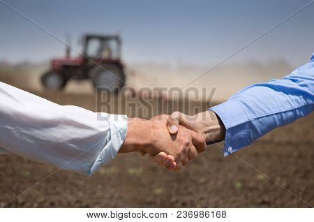 Two Businessmen Shaking Hands In Field With Tractor Working In Background. Agribusiness Concept