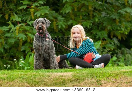 Pretty Young Blonde Girl Making Eye Contact While Sitting On Grass With Well Behaved Black Labradood