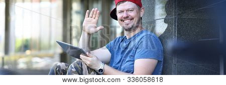 Portrait Of Stylish Man In Khaki Trousers, Blue T-shirt And Red Cap Waving With Hand Hello. Smiling 