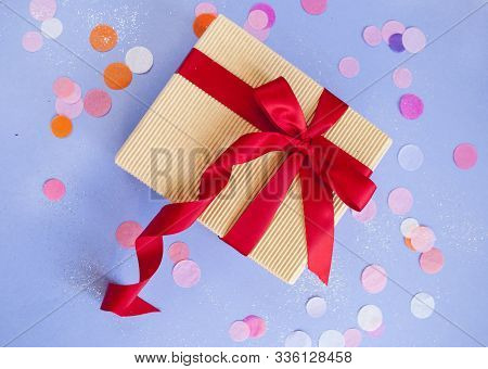 Giftbox Tied With Red Color Ribbon On Purple Background With Sparklets And Confetti. Flat Lay Style.
