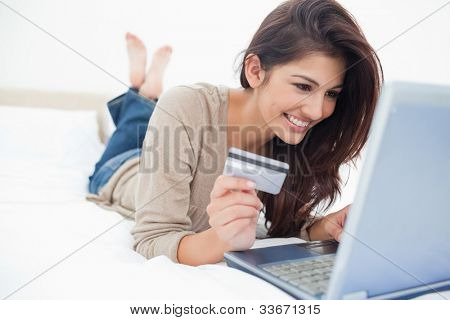 En smilende kvinde på hendes seng med laptop foran hende og kreditkort i hånden, med krydsede fødder.
