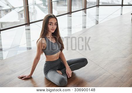 Photo From Above. Relaxed Cute Young Girl Sits On The Floor In Gym With Big Windows.