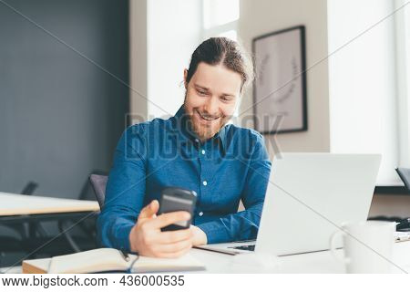 Smiling Young Male Employee In Casial Clothes Looking At Smartphone While Working On Laptop In Moder