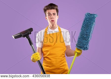 Dumbfound Male Homemaker In Apron And Gloves Holding Mop With Vacuum Cleaner While Looking At Camera