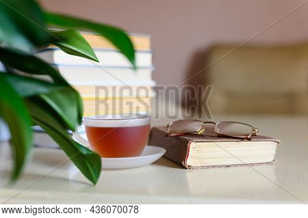 A Cup Of Tea Stands On A Closed Book Next To A Tall Stack Of Books On The Table At Home. Concept Of 