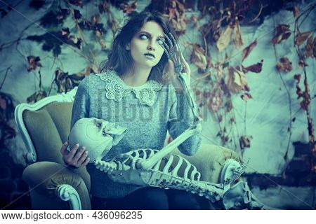 A pale ghost girl with dusty black hair and neat clothes sits in an old vintage armchair with a skeleton in her hands in the background of a ruined vintage wall. Halloween. Horror.