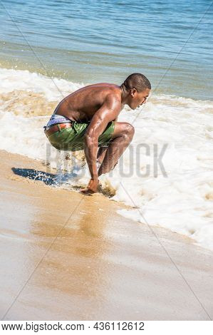 Bending Over, A Young Black Guy Is Squatting On The Beach, Playing With Waves