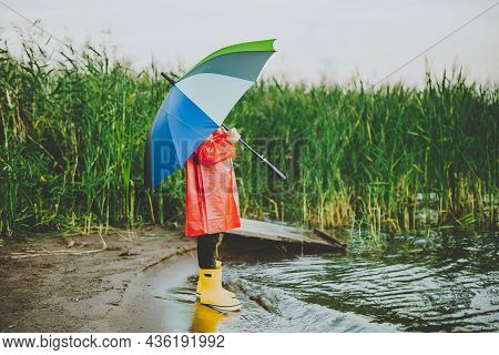 Boy In A Red Raincoat And Yellow Rubber Boots Stands At River Bank And Holding Rainbow Umbrella. Sch