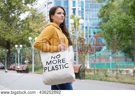 A Brunette Woman In An Orange Hoodie Walks With A Fabric Bag That Says No More Plastic In The City
