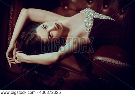 Adorable brunette woman pose lying on a red sofa in a gorgeous evening dress. Vintage beauty shot. Elegant evening style of the 30s-40s. 