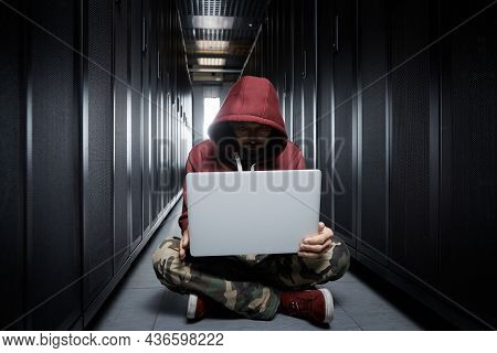 The Hacker In A Hood Sits With The Computer Among Servers, The Burglar, Face Covered With A Hood. St