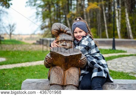 A Girl In The Park On A Bench Next To A Wooden Dwarf Sculpture, The Dwarf Reads A Book, Tervete