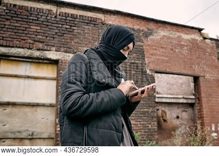 Young refugee female in hijab texting in mobile phone against wall of half ruined building