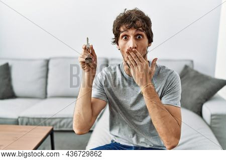 Young hispanic man holding keys of new home sitting on the sofa covering mouth with hand, shocked and afraid for mistake. surprised expression 