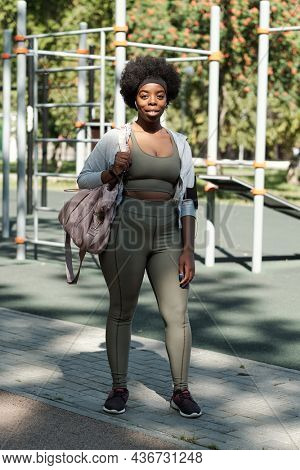 Young happy African sportswoman with handbag standing on sportsground before training