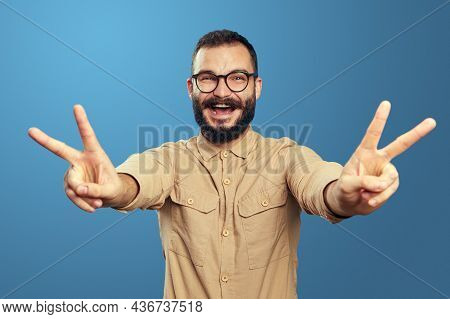 Excited Bearded Man Screams And Shows Victory Gesture With Both Hands, Wears Eyeglasses And Beige Sh
