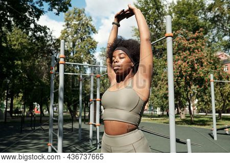 Young active sportswoman in tracksuit keeping her eyes closed and hands put together over head while exercising outdoors