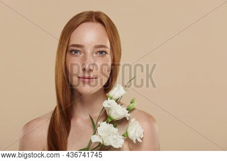 ?aucasian girl with naked shoulders holds eustoma flower. Cropped partial view of young beautiful red haired woman with freckles. Concept of natural female beauty. Isolated on beige background