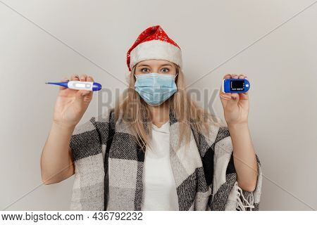 Woman In Santa Hat, Wrap And Protective Medical Mask Holds Thermometer And Puls Oximetr