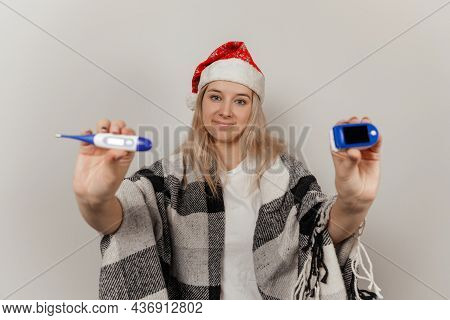 Woman In Santa Hat, Wrap And Protective Medical Mask Holds Thermometer And Puls Oximetr