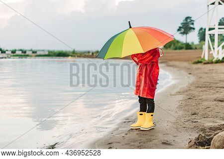 Boy In A Red Raincoat And Yellow Rubber Boots Stands At River Bank And Holding Rainbow Umbrella. Sch