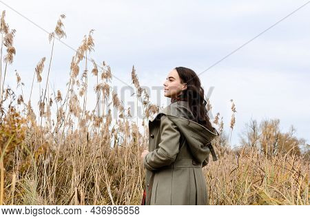 Young Dreamy Woman Wearing Grey Old Fashioned Coat Stands In The Dry Grass Reeds Field Looking Away.
