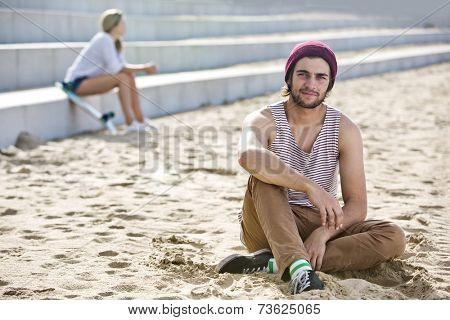 Profound looking dude, sitting in the sand on a beach with a woman sitting on concrete steps in the background.