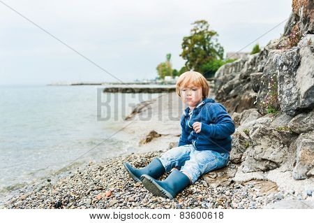 Cute toodler boy having fun outdoors, playing next to lake on a rainy day