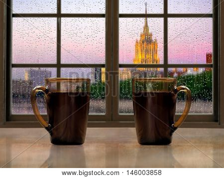 Two mugs with a hot drink - tea or coffee on the window sill of the window. During the early morning the city the rain. Concept - good morning alertness awakening together