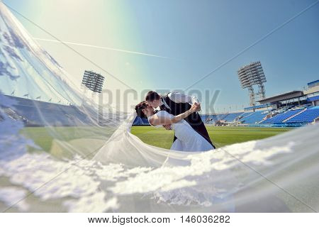 Wedding couple on the football stadium is hugging each other. Beautiful girl in white dress. Man in suit. Beauty bride with groom. Female and male portrait. Woman with lace veil. Lady and guy outdoors