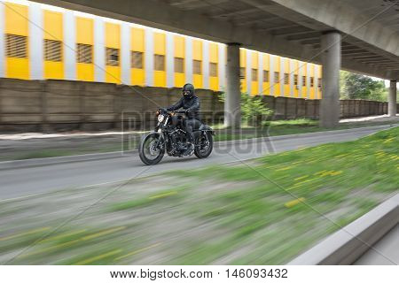 Guy in black helmet hurtling on the black motorbike on the road under the overpass. He wears a black leather jacket, black jeans, black shoes, black gloves and sunglasses. Outdoors. Horizontal.