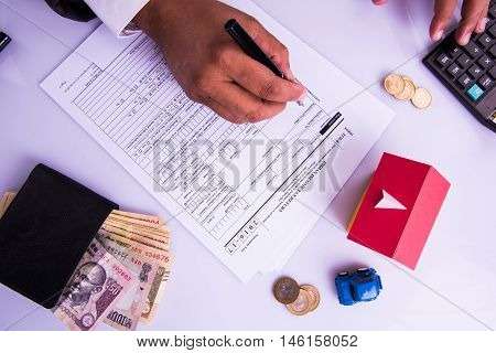 Indian man or accountant person filing Indian income tax returns form or ITR document showing indian currency, house model, toy car and calculator over white table top, selective focus