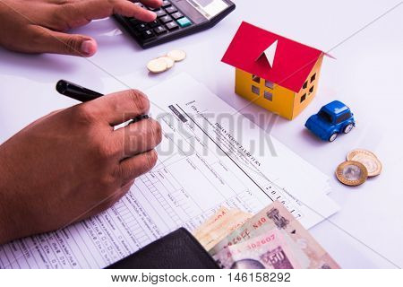 Indian man or accountant person filing Indian income tax returns form or ITR document showing indian currency, house model, toy car and calculator over white table top, selective focus