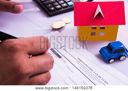 Indian man or accountant person filing Indian income tax returns form or ITR document showing indian currency, house model, toy car and calculator over white table top, selective focus