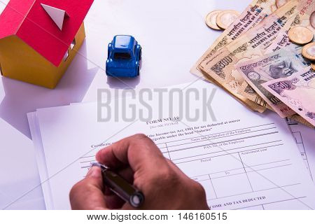 Indian man or accountant person filing Indian income tax returns form or ITR document showing indian currency, house model, toy car and calculator over white table top, selective focus