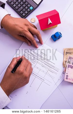 Indian man or accountant person filing Indian income tax returns form or ITR document showing indian currency, house model, toy car and calculator over white table top, selective focus