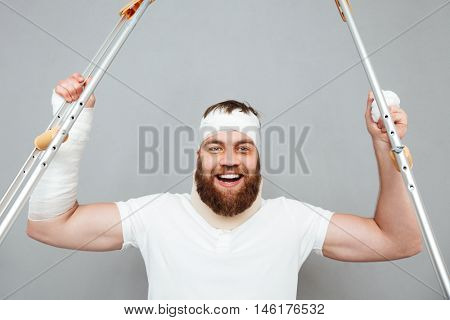 Cheerful bearded young man holdiing crutches and celebrating success over white background